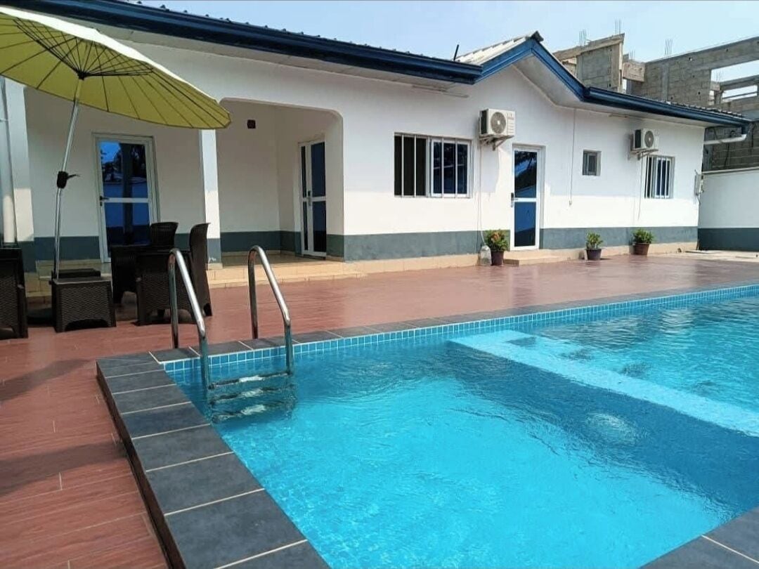 Piscina com borda revestida em azulejo, área coberta com cadeiras e um edifício com telhado azul e paredes brancas.