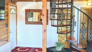A log cabin interior with a spiral staircase, a red patterned carpet, a leather sofa, and a small table with a plant. - Authentic Log Cabin w/ fire pit - 45 Mins to Yellowstone! (Island Park)