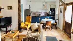 A cozy living room with a sofa, a yellow armchair, a TV, and a wooden coffee table. There is a dining area with a round table and chairs. A kitchen with blue cabinets and a patterned backsplash is visible in the background. - Charming cabin by a lake in the heart of the Landes d'Armagnac forest (Losse)