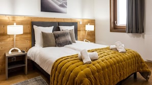A bedroom with a wooden headboard, two bedside lamps, a bed with white bedding and a mustard-colored quilt, a painting above the bed, and a window with curtains. - Apartment in Plagne 1800 With Ski-in/ski-out (La Plagne-Tarentaise)