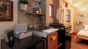 Una cocina compacta con pileta, cocina y una pequeña mesada. Hay una estantería con plantas y frascos, y un cuadro en la pared.
