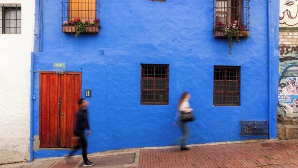 Un edificio azul con una puerta y ventanas rojas, una acera de ladrillo y un mural a la derecha.