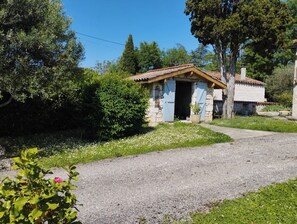 Situated in a lush green setting, this charming cottage features a small porch with shutters and a gravel driveway.