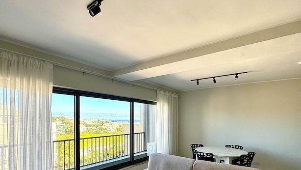 Una sala de estar moderna con un sofá color beige, una mesa de centro de mármol y una zona de comedor con una mesa blanca y sillas negras.