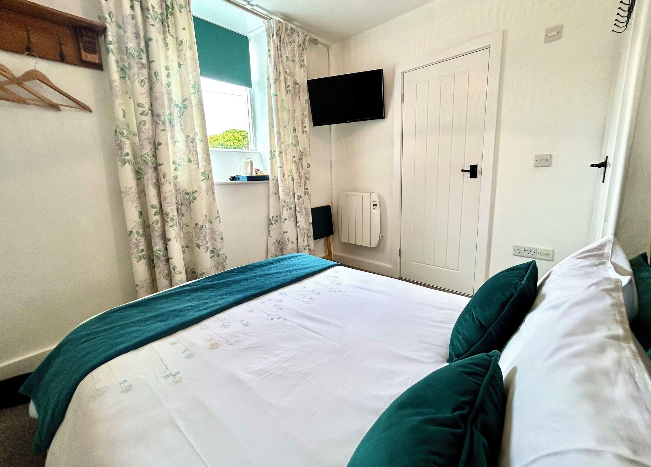 A bedroom with a bed, a window with curtains, a mounted TV, a radiator, and a door.