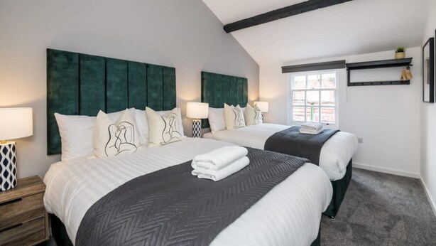 Une chambre avec deux lits, chacun doté d’une tête de lit verte, de draps blancs et de coussins décoratifs. Il y a une fenêtre donnant sur l’extérieur, ainsi qu’une étagère avec une plante accrochée au mur.