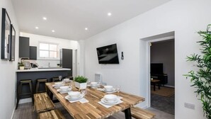 Un espace repas moderne avec une table en bois dressée pour quatre personnes, une cuisine en arrière-plan et un téléviseur fixé au mur.