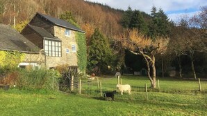 Una casa de piedra con techo inclinado, un patio cercado con ovejas y un árbol en primer plano.