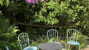 Un jardín con una mesa y sillas de metal, rodeado de follaje denso y una cerca de alambre.