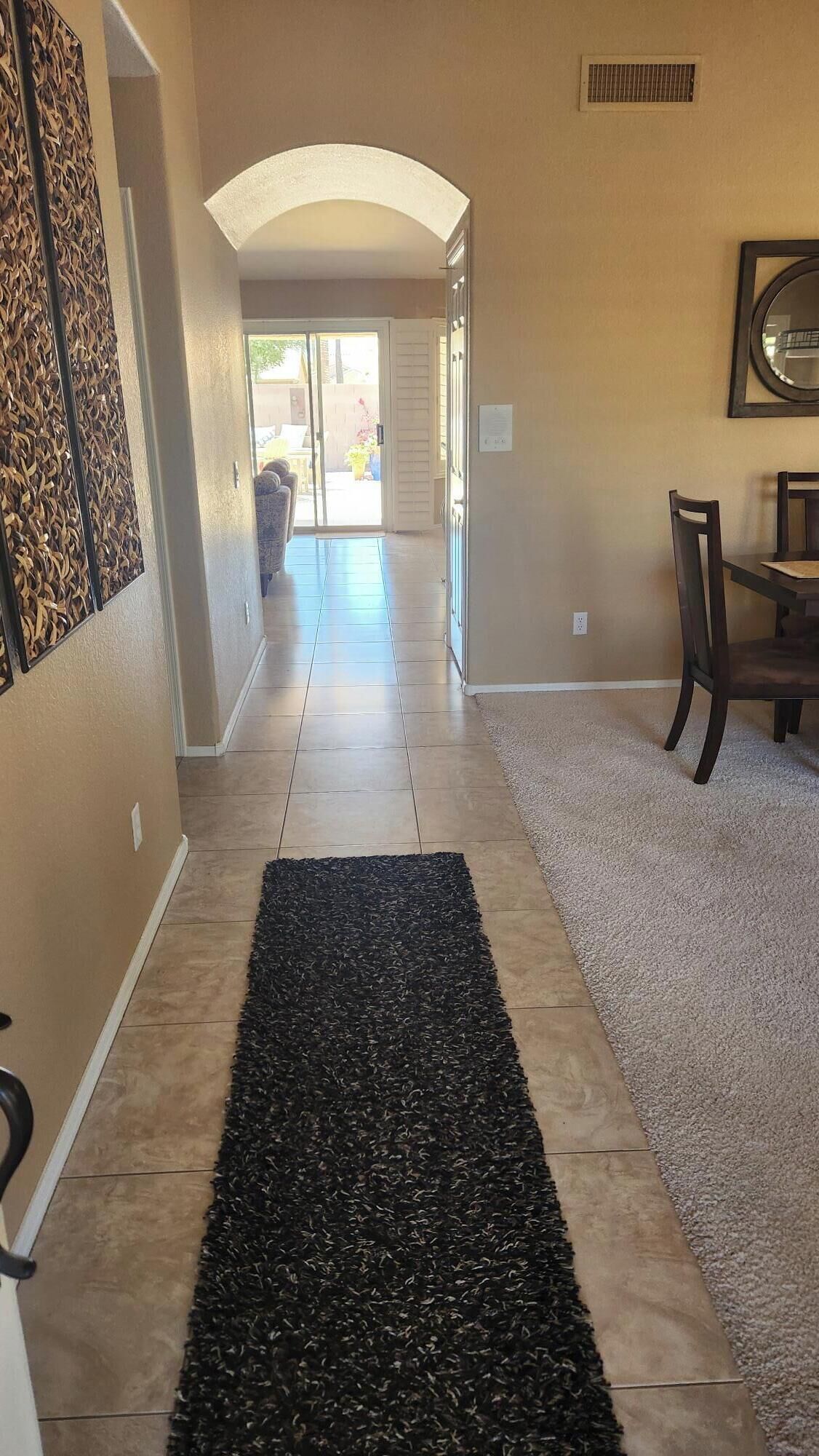 A hallway with a black rug, beige walls, a wooden chair, and a dining table.