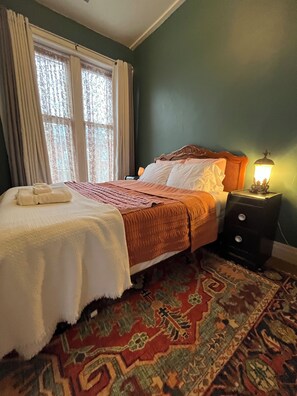 Un dormitorio con cama, mesita de noche, lámpara y ventana con cortinas.