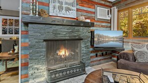 Una sala de estar acogedora con chimenea de piedra, televisor montado y ventana con vistas a árboles.