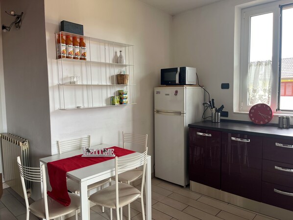 Une cuisine de taille réduite avec un réfrigérateur blanc, un four à micro-ondes placé sur le dessus, et une table à manger recouverte d’une nappe rouge.