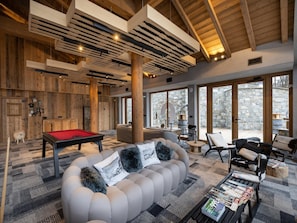 A spacious living area with a pool table, a large sofa with decorative pillows, and a coffee table with magazines. The room features wooden beams on the ceiling and large windows offering a view of the outdoors. - Apartment in Megeve Near Fornelet Cable Car (Megeve)