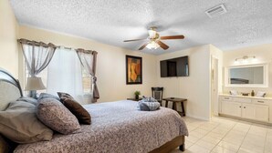A bedroom with a bed, curtains, a ceiling fan, a TV, a small table with a plant, and a bathroom with a mirror and cabinets. - Private San Antonio Getaway Near SeaWorld! (San Antonio)