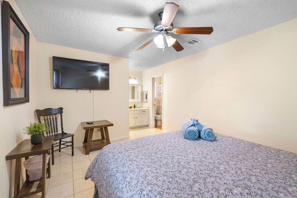 Une chambre à coucher avec un lit, un ventilateur de plafond, une télévision, un bureau, une chaise et une salle de bain visible à travers une porte ouverte.
