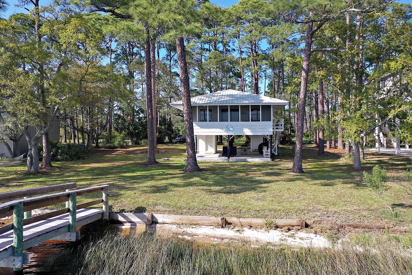 A white building with a covered patio, surrounded by tall trees and a wooden bridge.