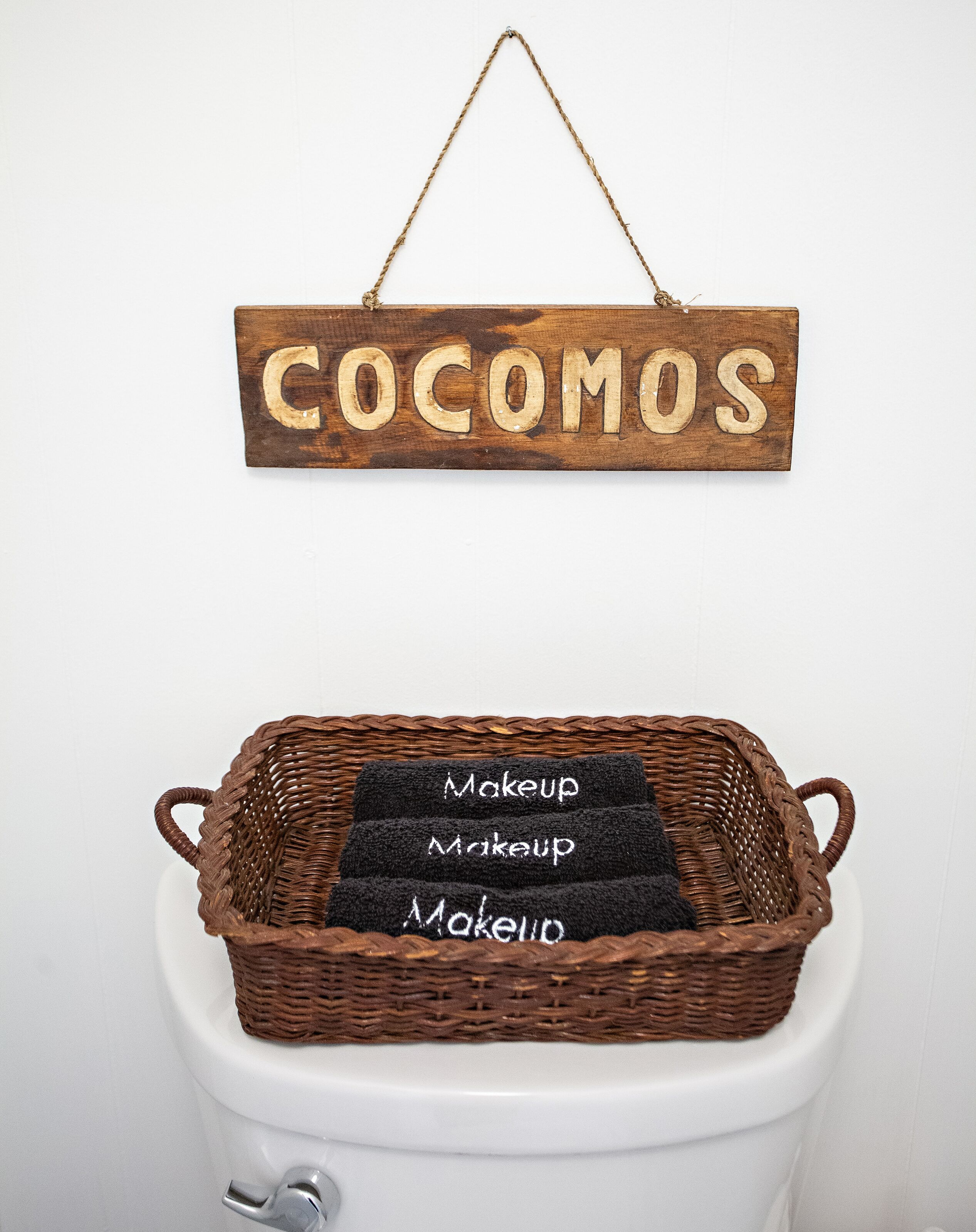 A wooden sign with the word 'COCOMOS' hanging above a wicker basket containing towels labeled 'Makeup' on a toilet.
