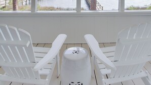 A covered outdoor area with two white Adirondack chairs and a white trash can, overlooking a body of water with a dock and trees.