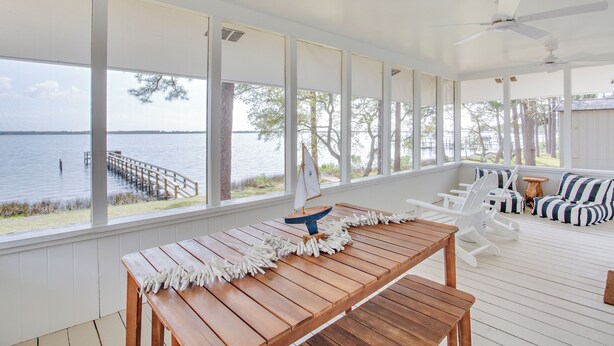 A covered outdoor area with a wooden deck, white furniture, and a view of the water.