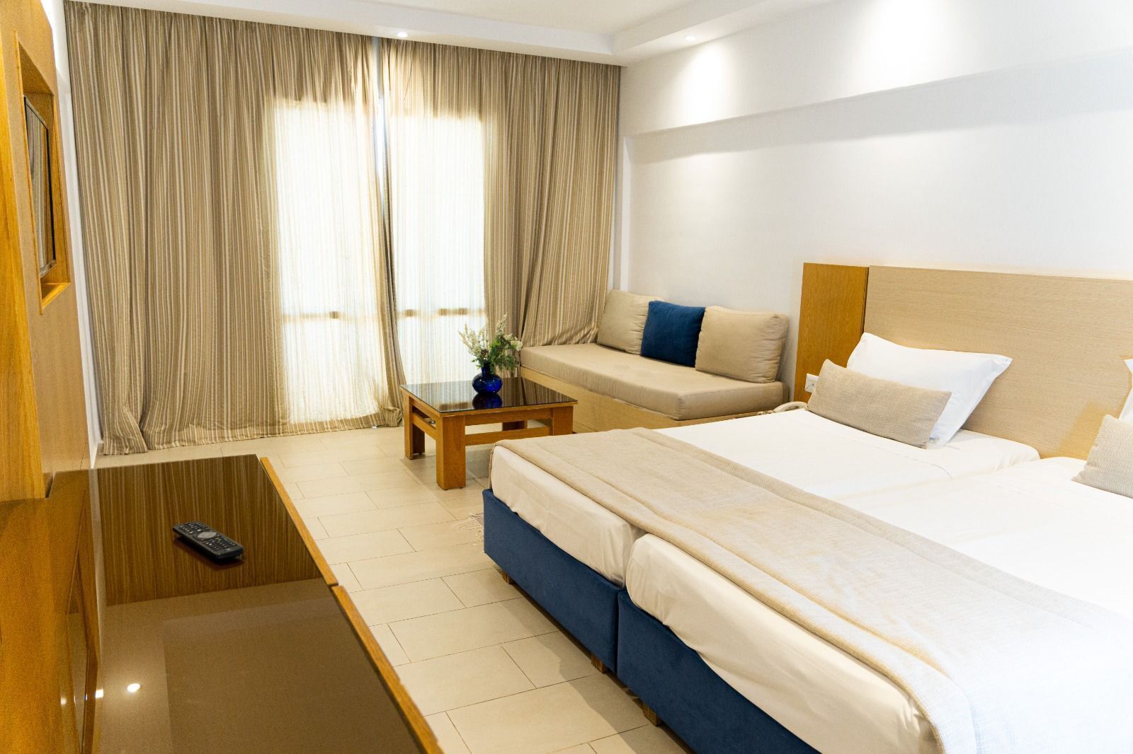 A hotel room with a large bed, a sofa, a small table with a vase, and a remote control on the floor.