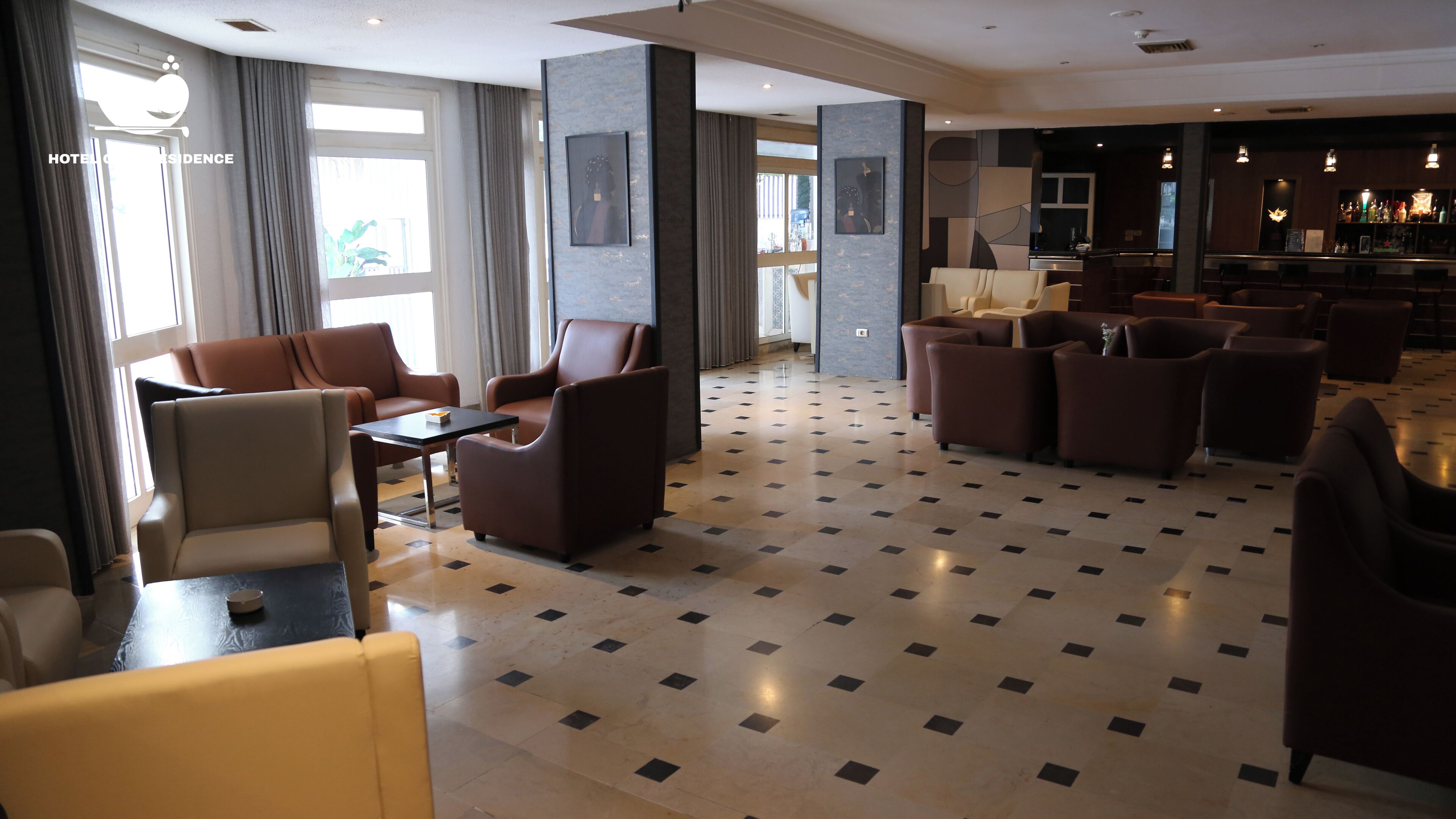 Un hall d’hôtel avec un sol à carreaux, des canapés en cuir, un coin bar et de grandes fenêtres avec des rideaux.
