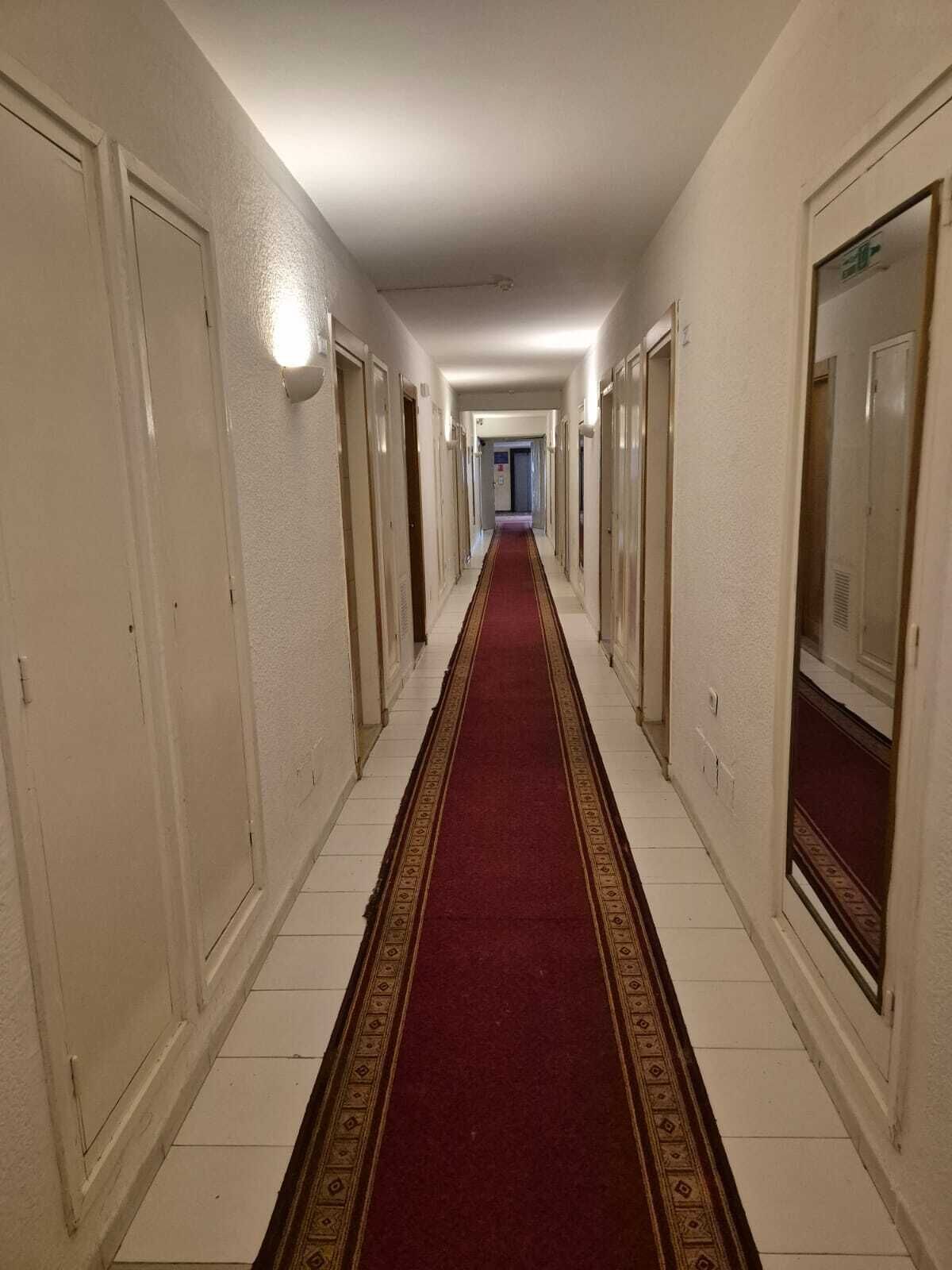 A long, narrow hallway with white walls and doors on both sides, leading to a room at the end.