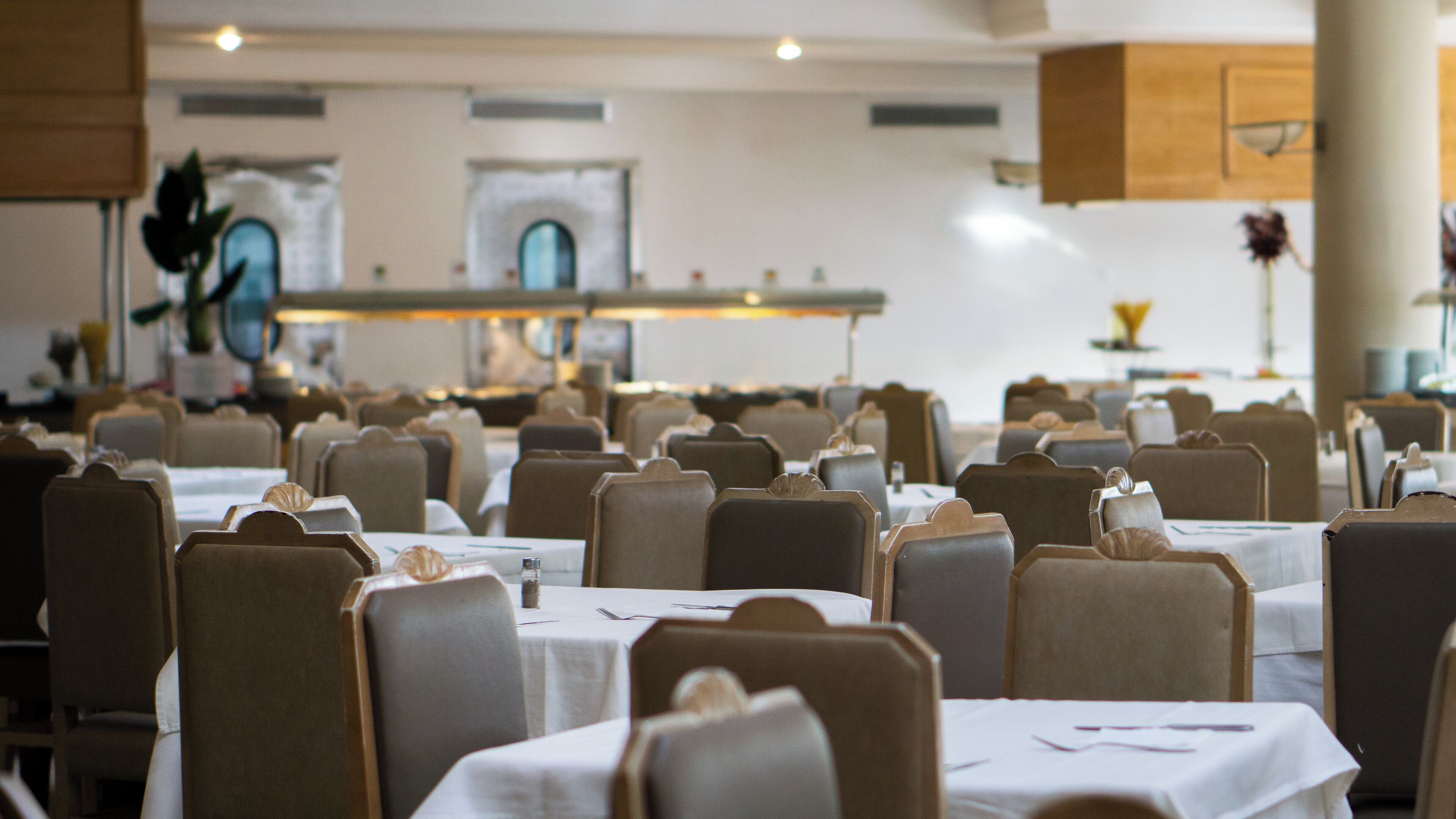 Ein Restaurant mit mehreren Tischen und Stühlen, einem Küchenbereich im Hintergrund und einer neutralen Farbgestaltung.