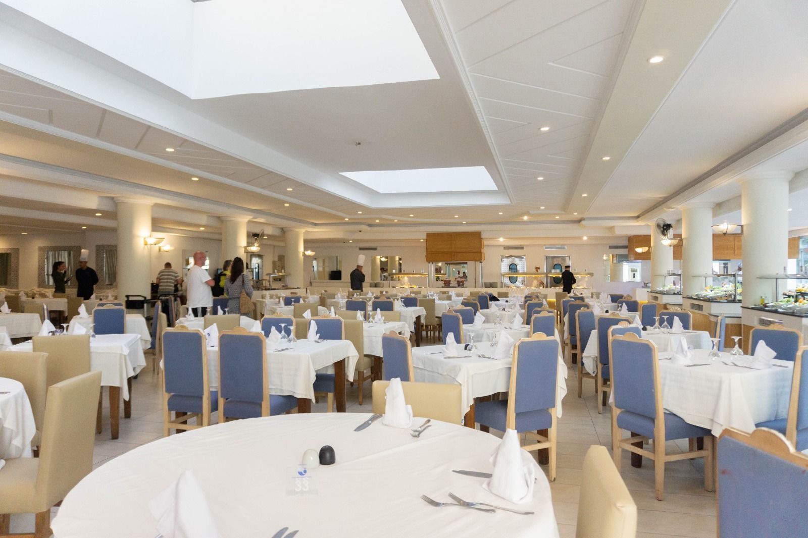 Un vaste espace dédié à la salle à manger, avec de nombreuses tables et chaises, un plafond haut doté de lucarnes et une cuisine centrale.
