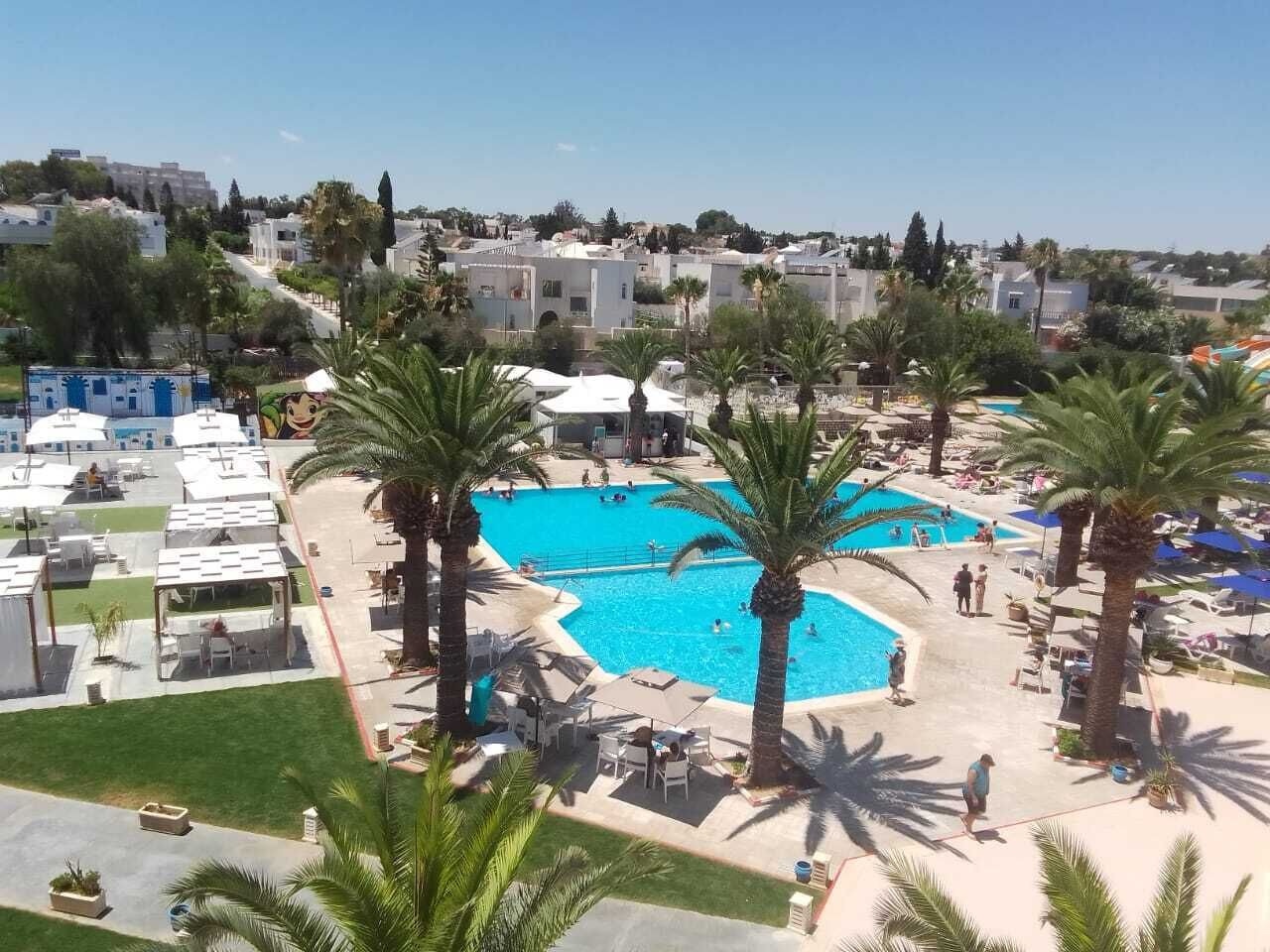 Ein Resort mit einem zentral gelegenen Swimmingpool, umgeben von Palmen und Liegestühlen.