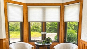 Ein Eckzimmer mit zwei Sesseln, einem kleinen Tisch und Blick auf Grün durch große Fenster mit Holzrahmen.