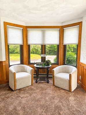Une pièce d’angle comprenant deux fauteuils, une petite table et une vue sur la verdure grâce à de grandes fenêtres à cadre en bois.