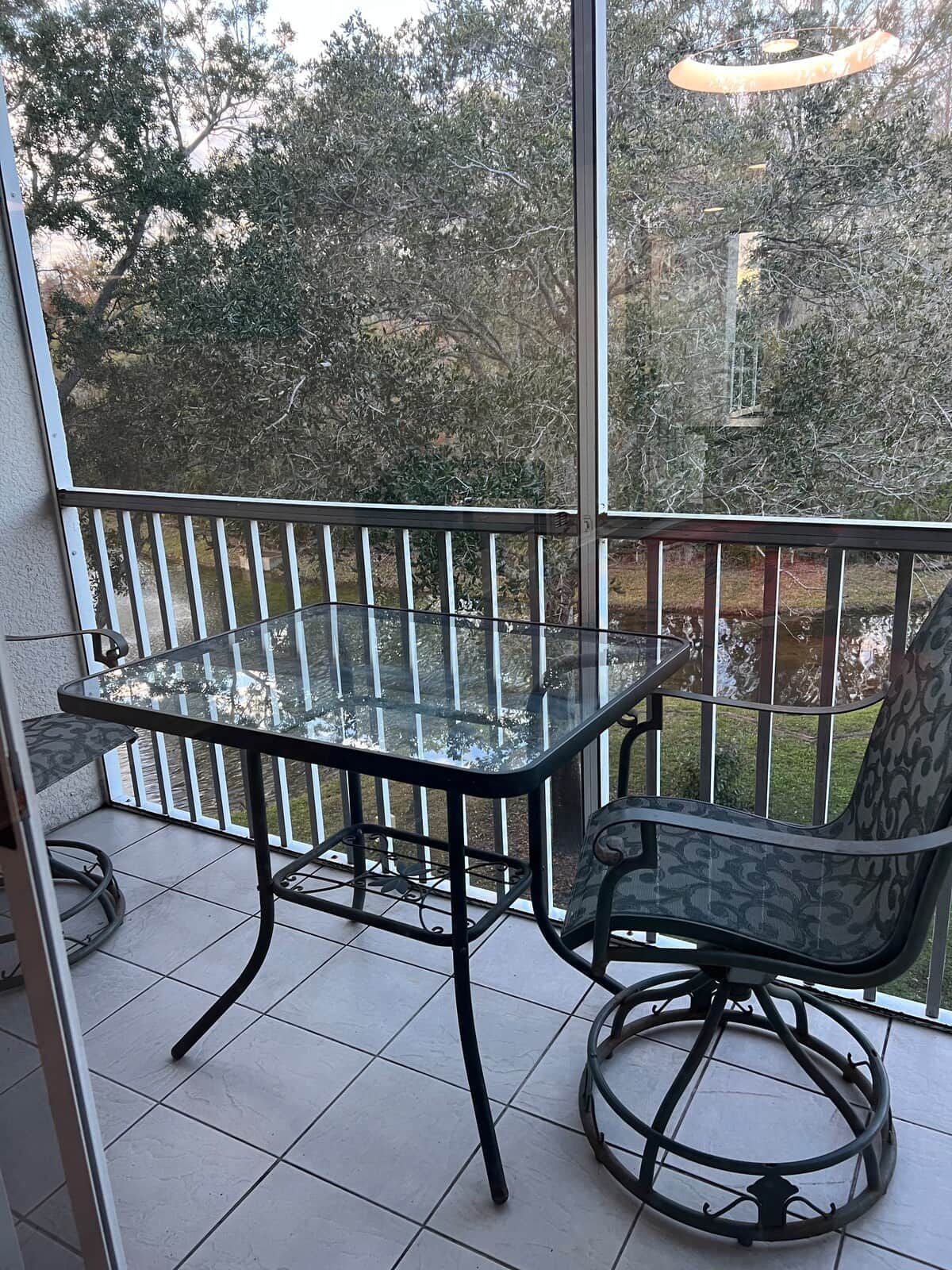 Una mesa de vidrio con una silla en un balcón con barandilla, con vistas a un jardín con árboles.