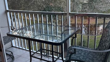 Una mesa de vidrio con una silla en un balcón con barandilla, con vistas a un jardín con árboles.