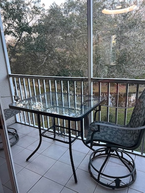 Ein Tisch mit Glasplatte und ein Stuhl auf einem Balkon mit Geländer, mit Blick auf einen Garten mit Bäumen.