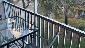 Un balcon avec une table et des chaises en verre, donnant sur une étendue d’eau et des arbres.
