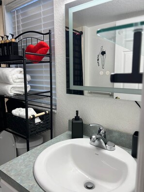 Une salle de bain avec un lavabo blanc, un porte-serviettes noir avec des serviettes, un miroir et un distributeur de savon fixé au mur.