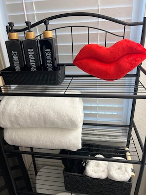 Un estante metálico con artículos de aseo, un cojín rojo con forma de labio y toallas dobladas.