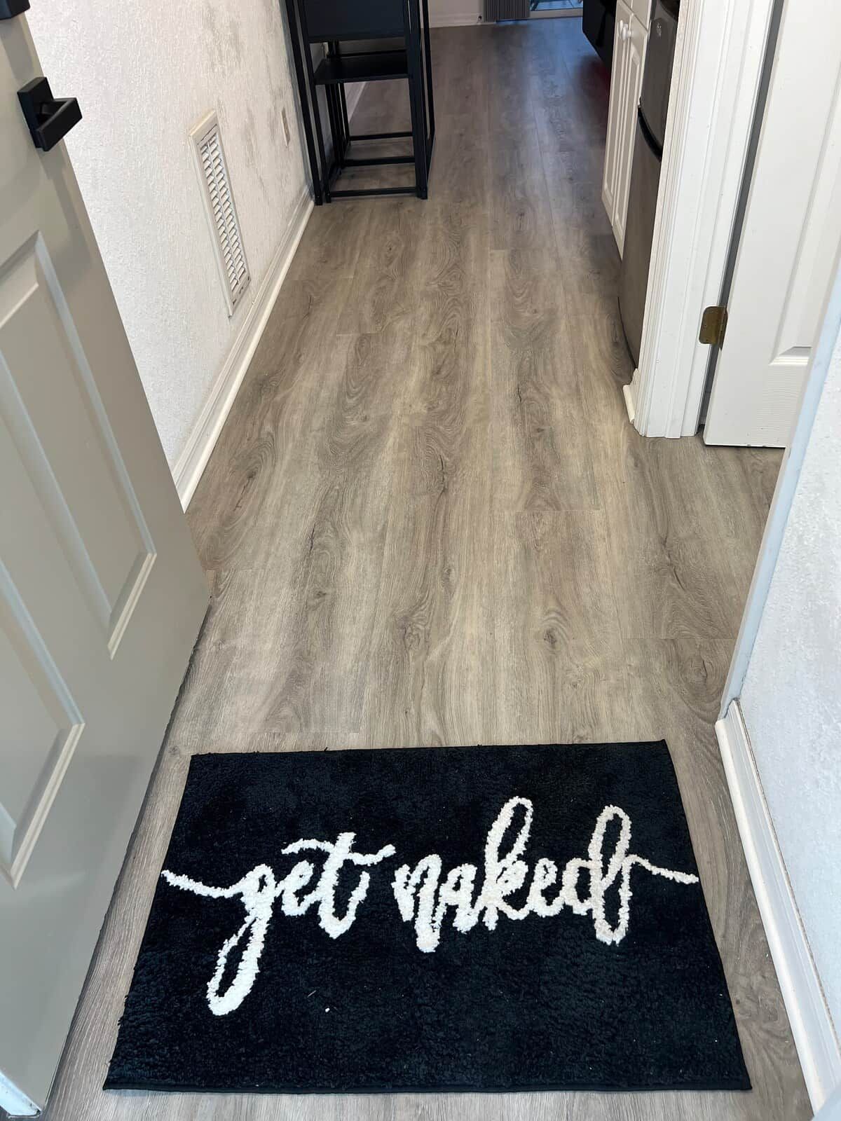 Een gang met een zwarte deurmat waarop 'get naked' staat en een houten vloer.