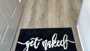 Een gang met een zwarte deurmat waarop 'get naked' staat en een houten vloer.