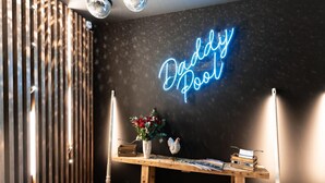 Una habitación con un letrero de neón que dice "Buddy Pool", una mesa de madera con libros y un jarrón de flores, y una pala de nieve en el suelo.