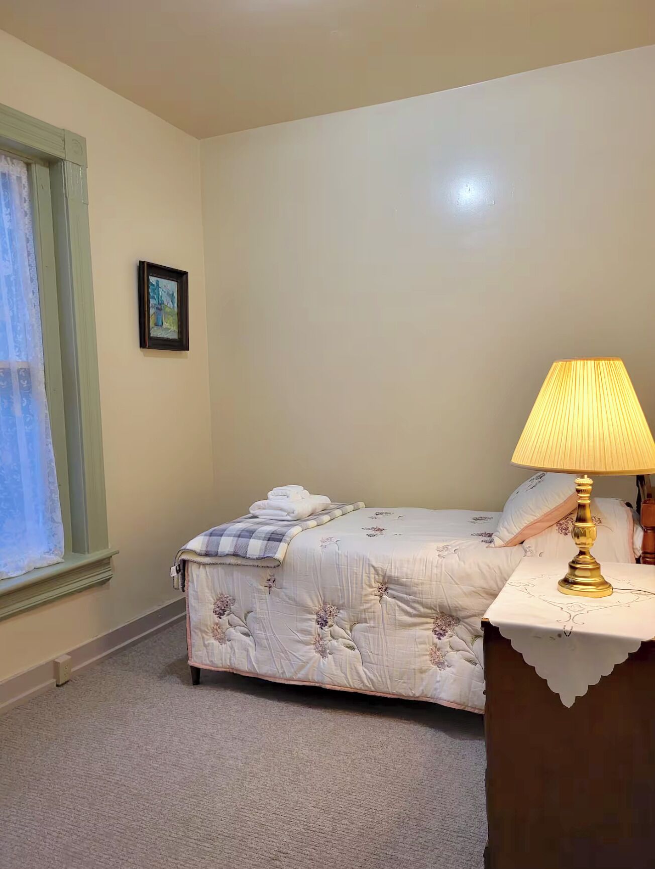 A bedroom with a bed, a bedside table with a lamp, a window with curtains, and a framed picture on the wall.