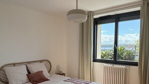 A bedroom with a large bed, a window with a view of the sea, a ceiling fan, and a radiator.