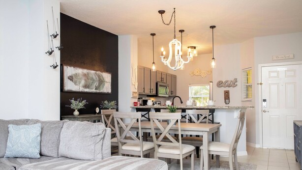 A modern living room with a gray sofa, wooden dining table, and chairs. A chandelier hangs above the table, and a kitchen is visible in the background.
