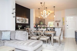 A modern living room with a gray sofa, wooden dining table, and chairs. A chandelier hangs above the table, and a kitchen is visible in the background.