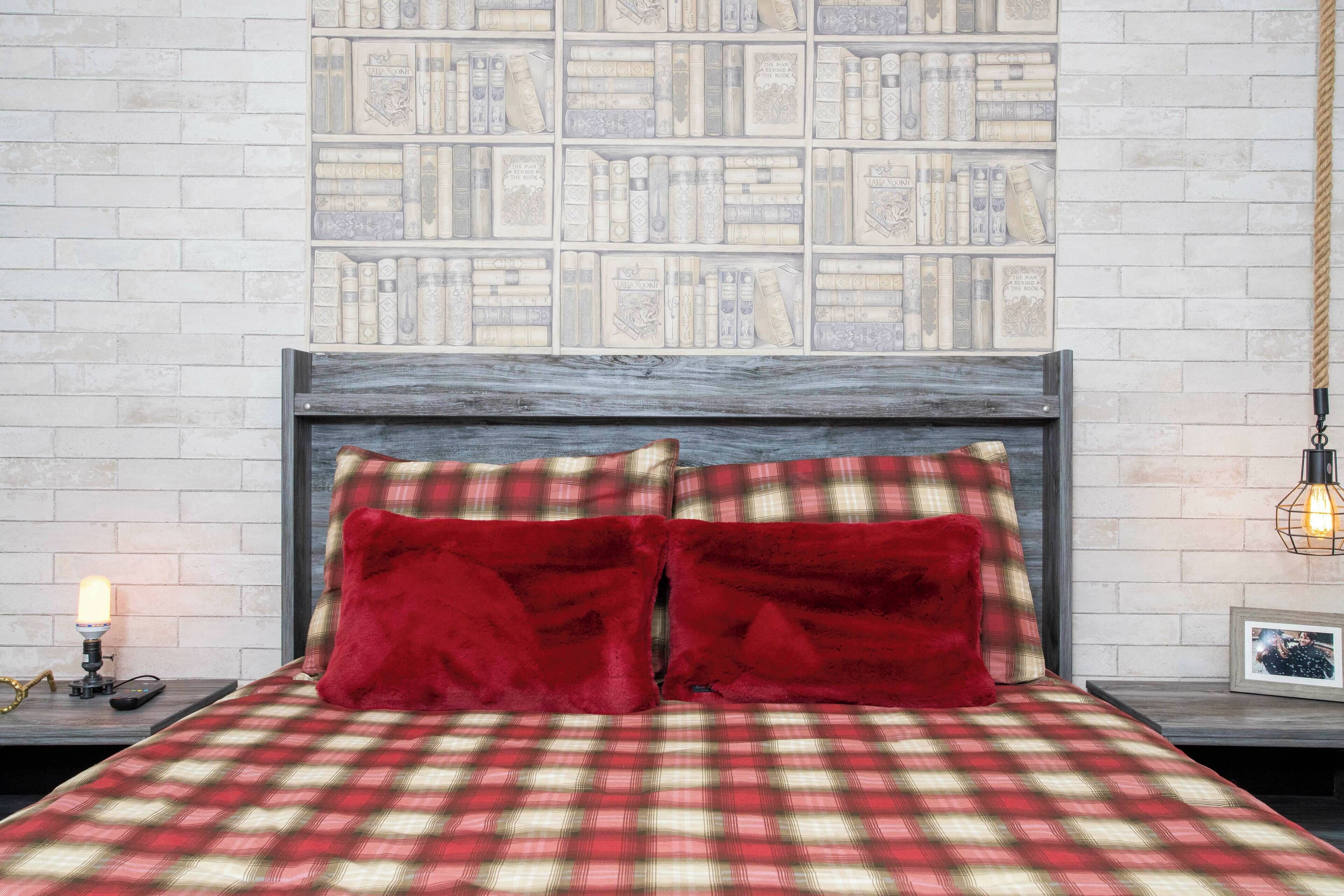 Uma cama com colcha xadrez e duas almofadas vermelhas, uma parede de tijolos com uma estante, e uma mesa de cabeceira com uma luminária e uma foto emoldurada.