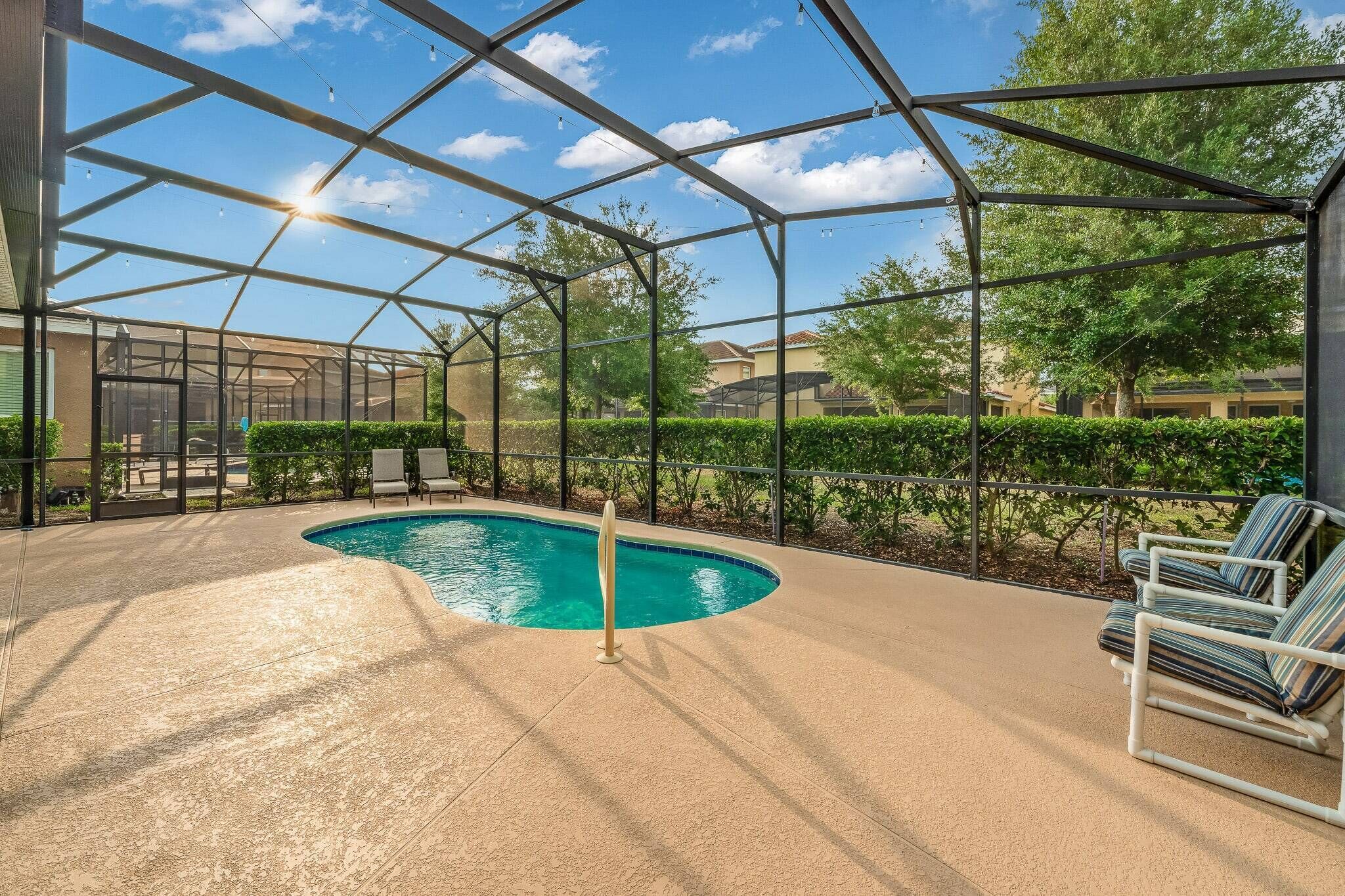 A covered outdoor swimming pool with a glass enclosure, surrounded by a landscaped area with hedges and trees, and a few lounge chairs.