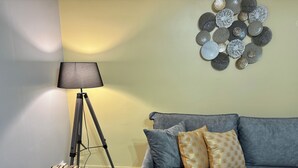 Ein Zimmer mit grauem Sofa, zwei gemusterten Kissen, einer Stehlampe, einer Holzbank und einem Wandbild.