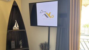Ein Wohnzimmer mit an der Wand befestigtem Fernseher, einem dunklen Holzregal, einem kleinen Holzschemel und einer Vase mit Trockenpflanzen.