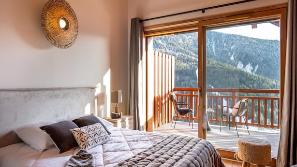 Een slaapkamer met een groot bed, uitzicht op een balkon met stoelen en een berglandschap.